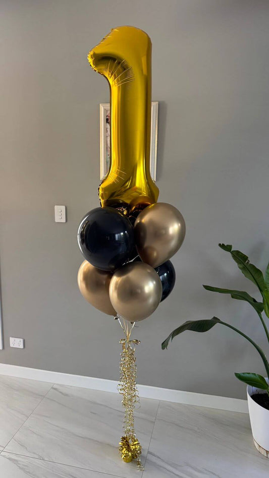 Gold, black, and gold balloons shaped like the number '1' against a neutral wall.